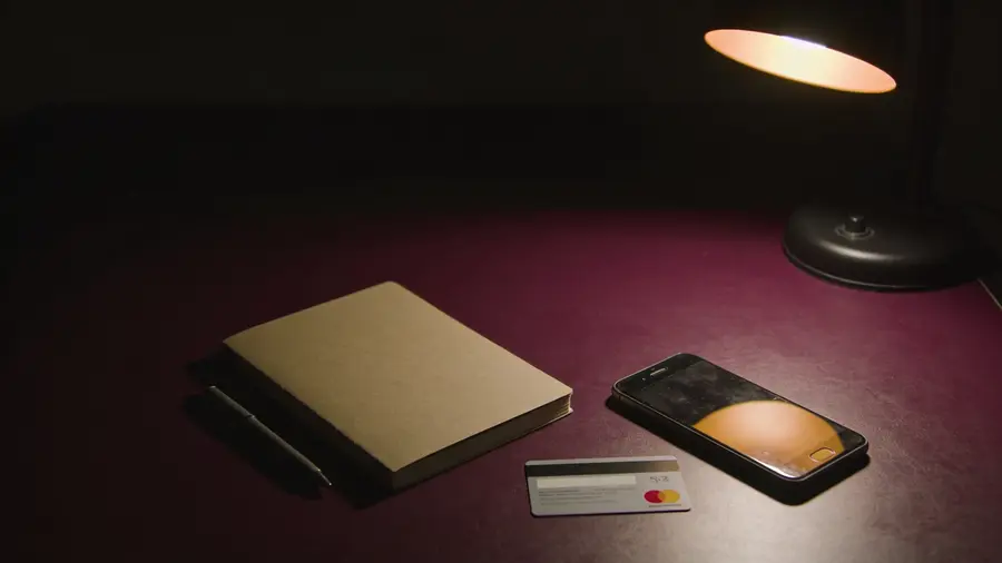 A notebook, pen and bank card on a dark desk beside a dimmed phone